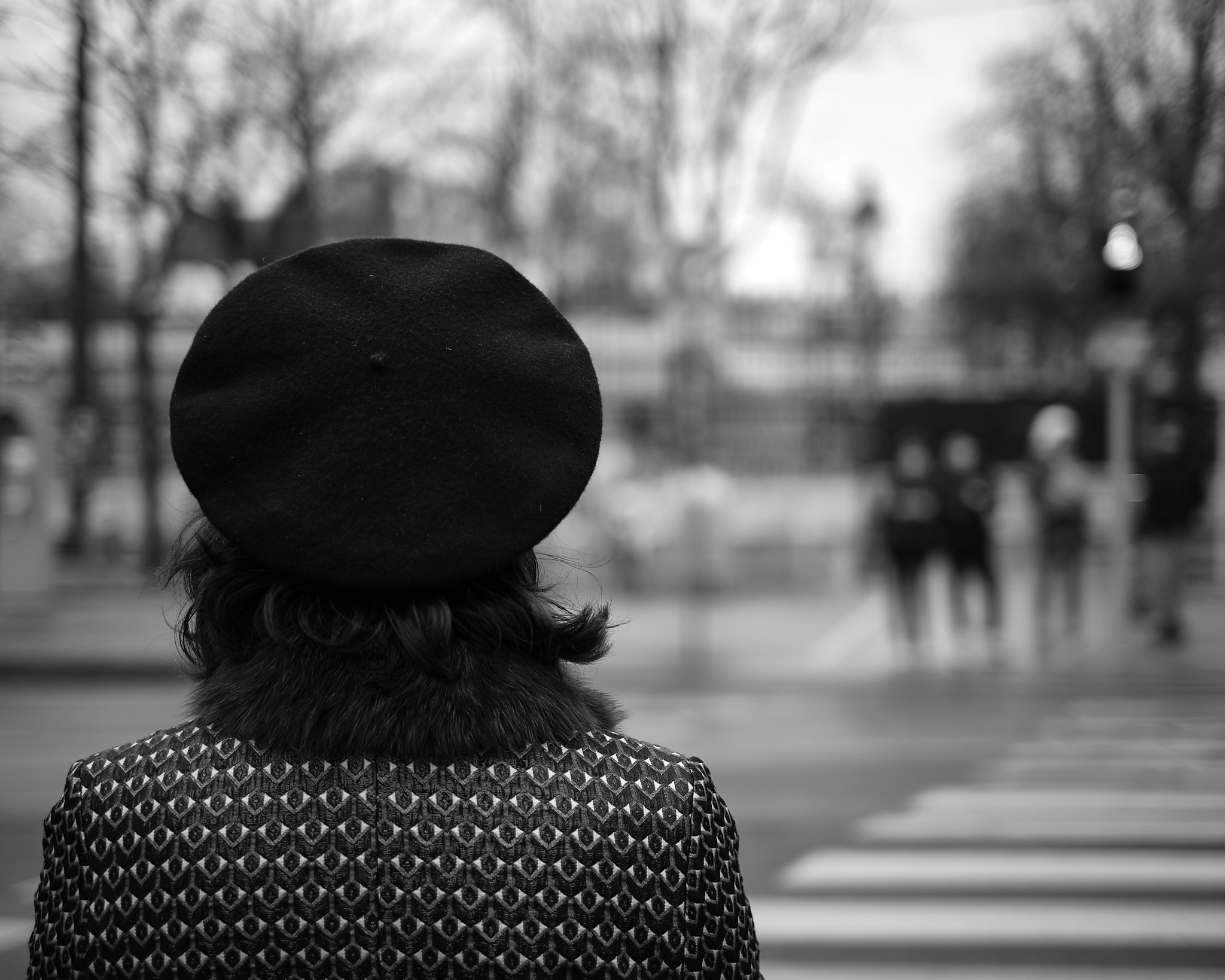 A person with shoulder-length hair, wearing a beret and a patterned coat, stands at a crosswalk. The background is blurred, with a few figures in the distance, and bare trees line the street.